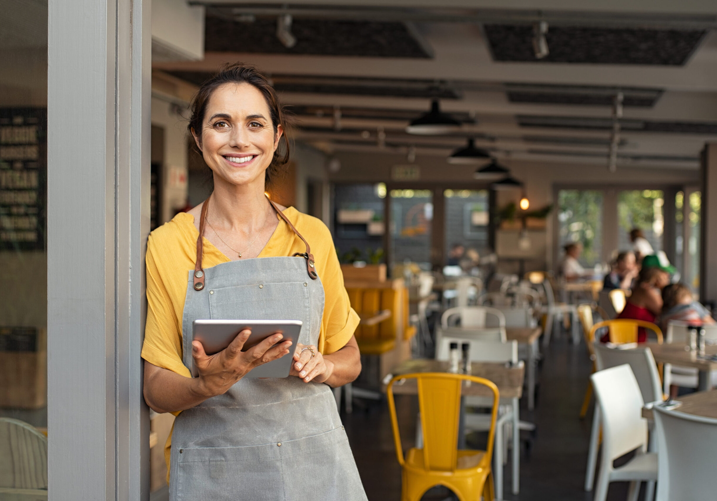 Small business owner at entrance looking at camera Portrait of happy woman standing at doorway of her store holding digital tablet. Cheerful mature waitress waiting for clients at coffee shop. Successful small business owner in casual clothing and grey apron standing at entrance and looking at camera.
